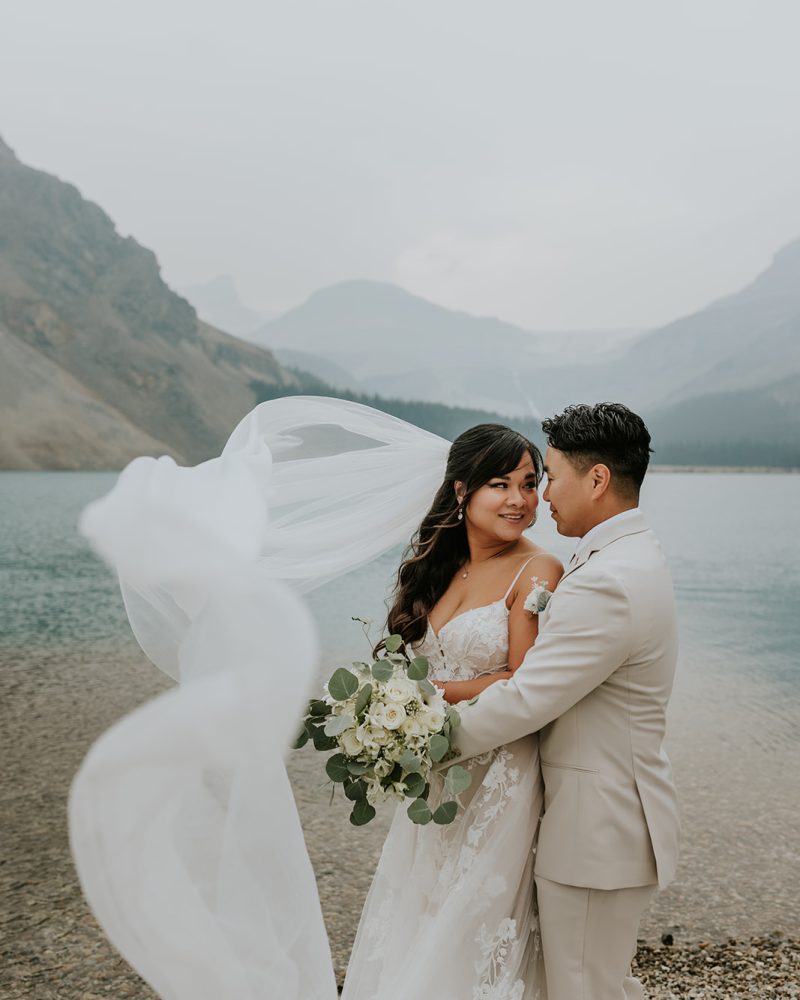 A bride and groom embrace by a mountain lake in Canmore, her veil flowing in the wind. The bride holds a bouquet of white flowers and greenery, with misty mountains rising in the background—a perfect elopement setting.
