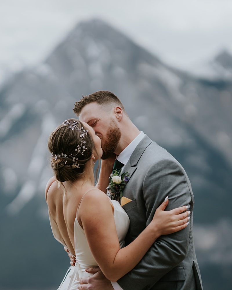 A bride and groom embrace and kiss outdoors at their Canmore wedding, with misty, snow-capped mountains and cloudy skies in the background. The bride wears a white dress and hairpiece, while the groom wears a gray suit.