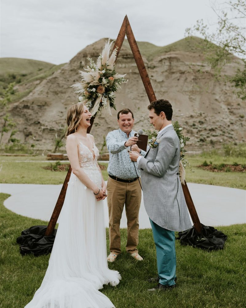 A bride and groom stand outdoors on grass in front of a wooden triangular arch with flowers, as an officiant holds a book. The groom reads vows while the bride smiles, with hills and their wedding photographer capturing the moment in the background.