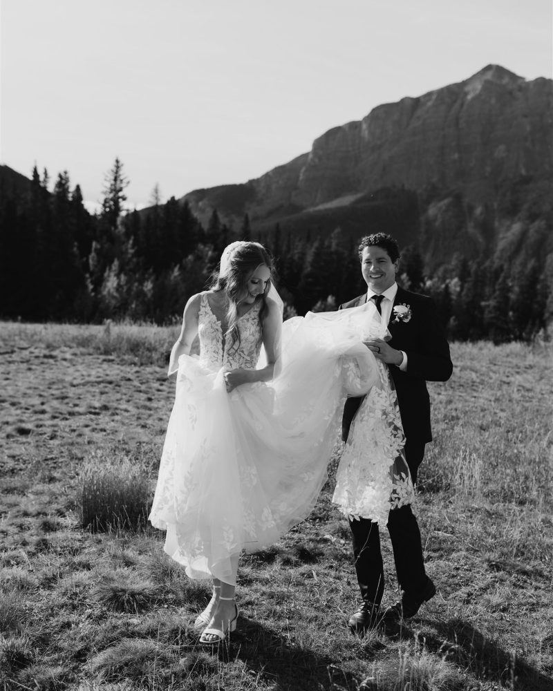 A bride and groom walk together outdoors in a grassy field with mountains in the background. The Canmore wedding photographer captures the groom lifting the bride’s dress as they smile, enjoying a sunny day in this black and white image.