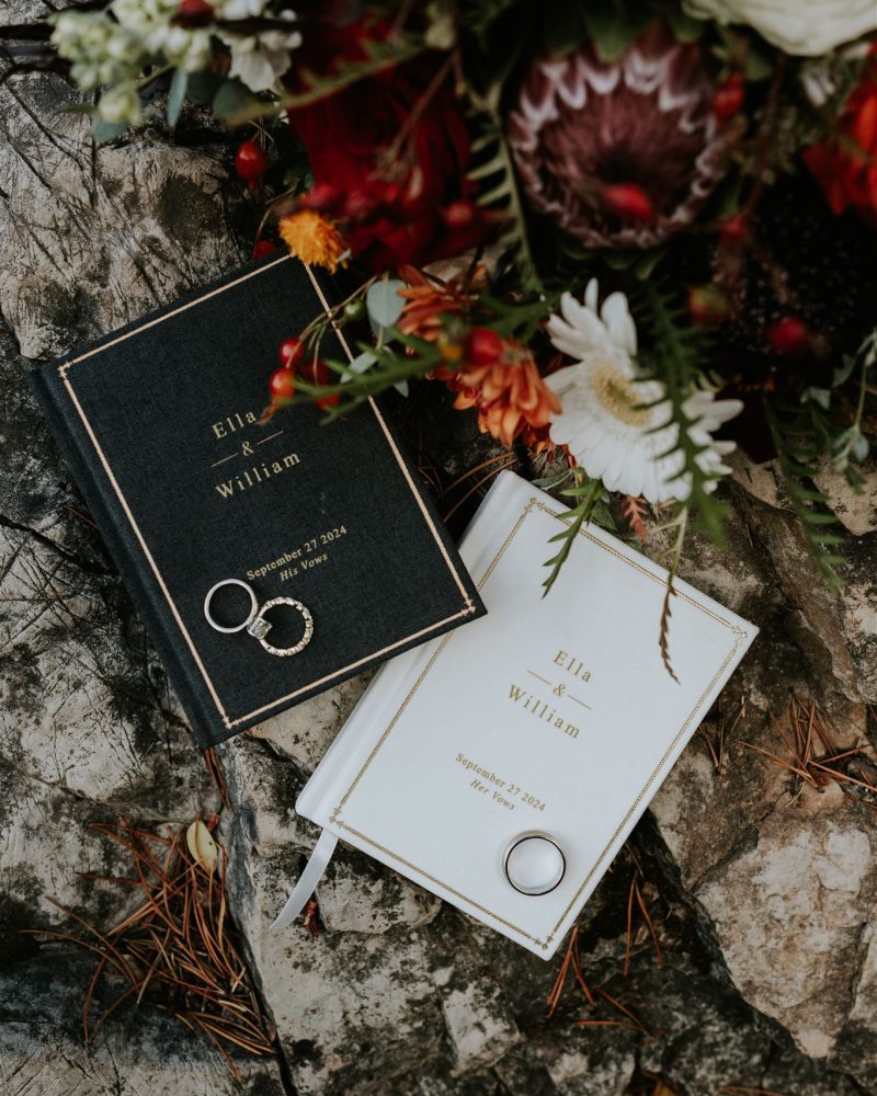 Two vow books, one black and one white, with Ella & William and a wedding date on the covers, lie on a rocky surface with pine needles. A Banff elopement photographer captured three rings and a bouquet of red and white flowers nearby.