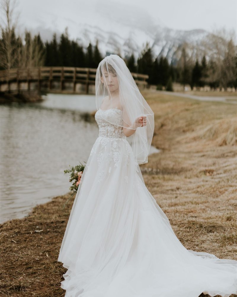 A bride in a white wedding gown and veil stands by a lakeshore near Banff, holding a bouquet. A wooden bridge, bare trees, and snow-capped mountains form the backdrop beneath a cloudy sky.