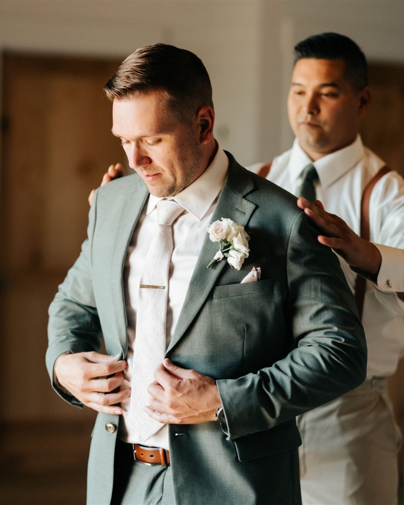A man in a suit adjusts his jacket while another, in a dress shirt and suspenders, stands behind helping—preparing for a formal elopement in Canmore. The softly lit room suggests a photographer is capturing this special moment.