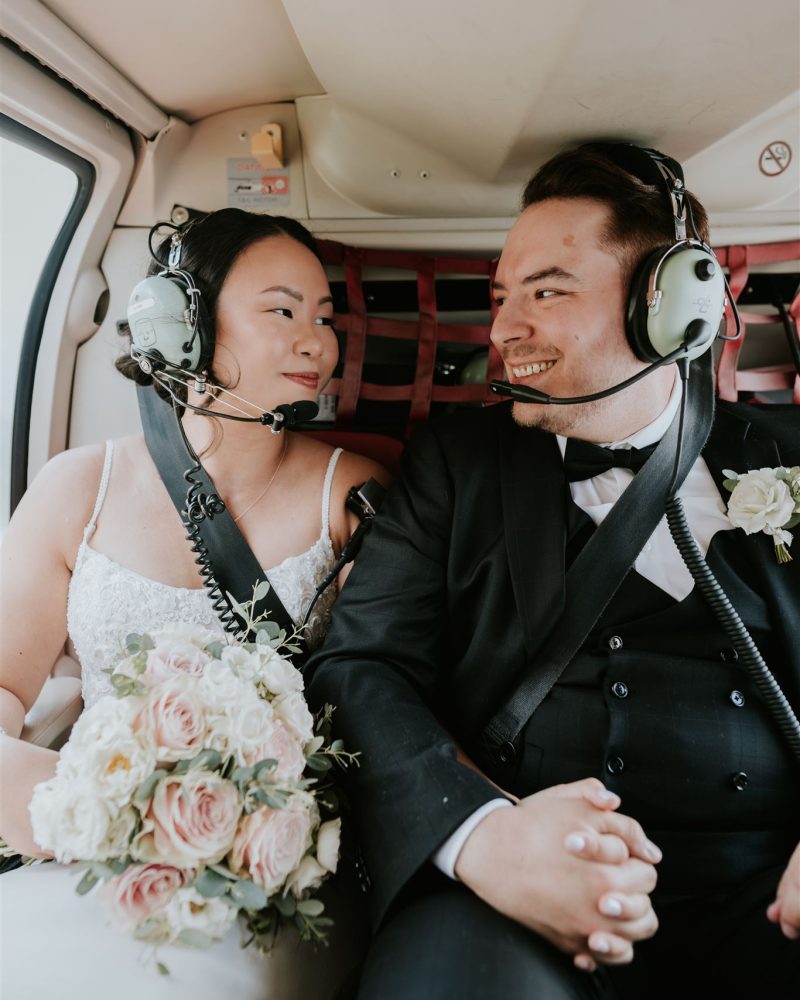 A bride and groom wearing headsets sit inside a helicopter over Banff, holding hands and smiling at each other. The bride, clutching a bouquet of pale pink and white roses, looks thrilled as their wedding photographer captures the joyful moment.
