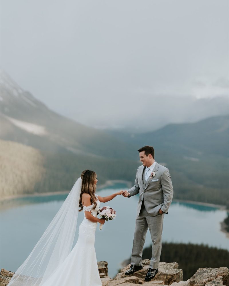 A bride in a white gown and veil holds a bouquet as she stands on a rocky ledge, reaching out to her groom in a light gray suit. Behind them, Canmore’s scenic lake and forested mountains create the perfect wedding backdrop under a cloudy sky.