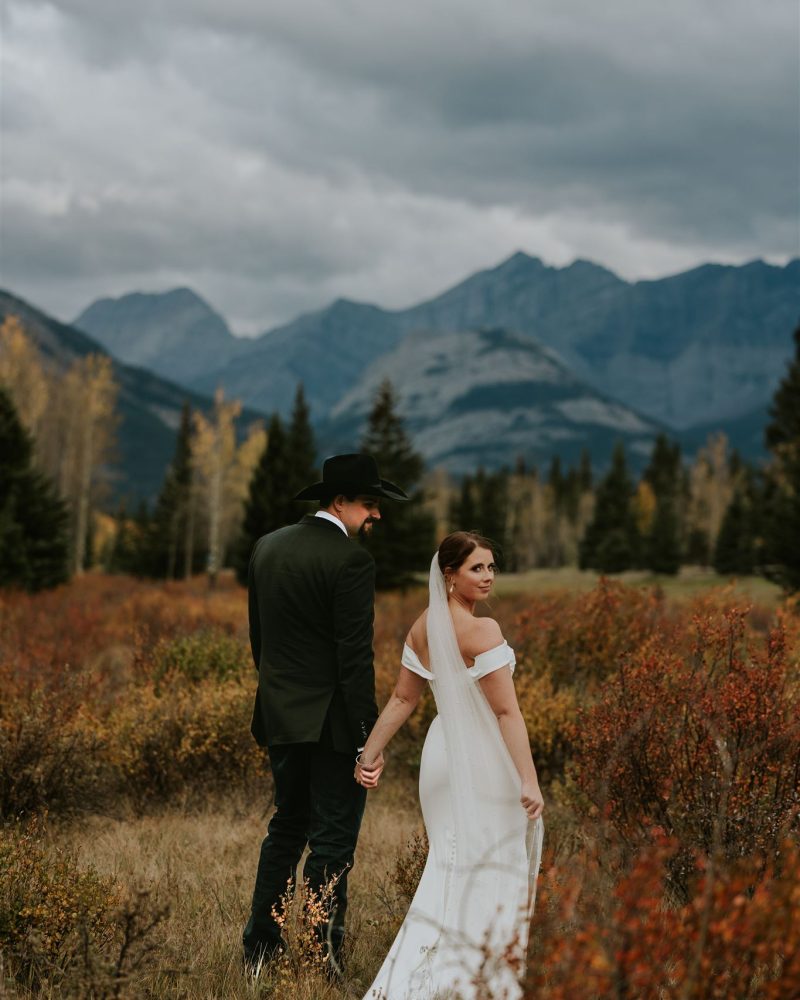 A bride and groom stand in an autumn meadow near Canmore, holding hands with mountains behind them. Captured by a photographer, this elopement features the bride in a white dress and veil, the groom in a dark suit and hat under an overcast sky.