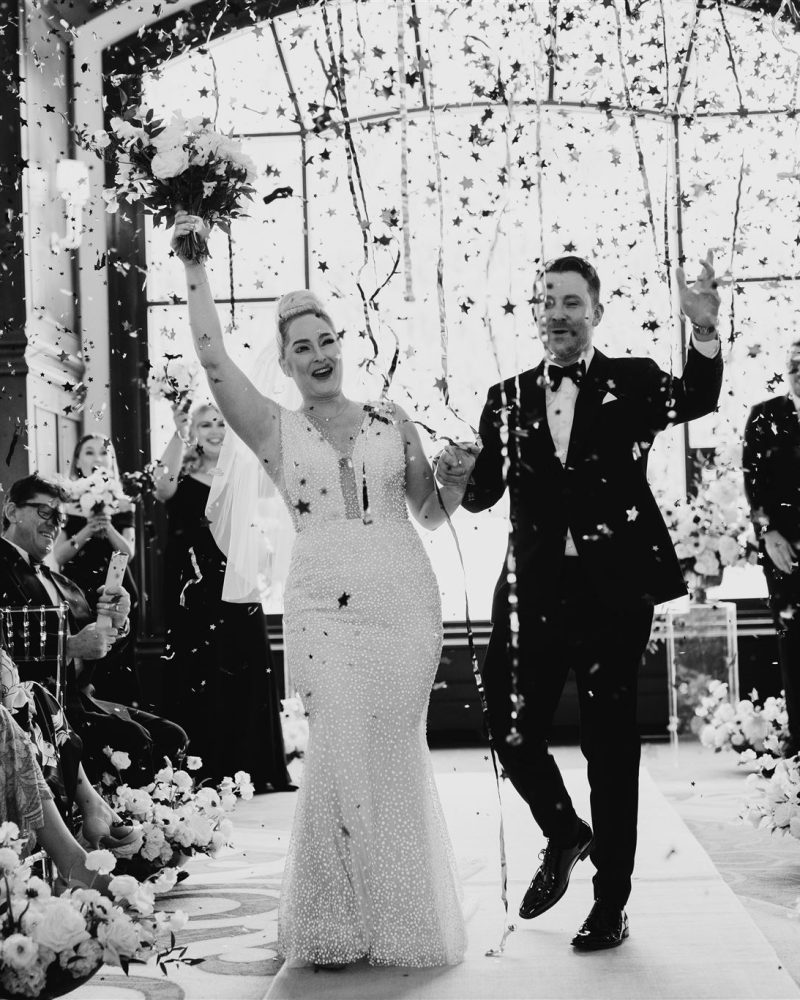 A newlywed couple walks down the aisle at their Canmore wedding, smiling and holding hands, as guests cheer and toss confetti. The bride's white dress and bouquet shine beside the groom’s tuxedo in this joyful and celebratory scene.