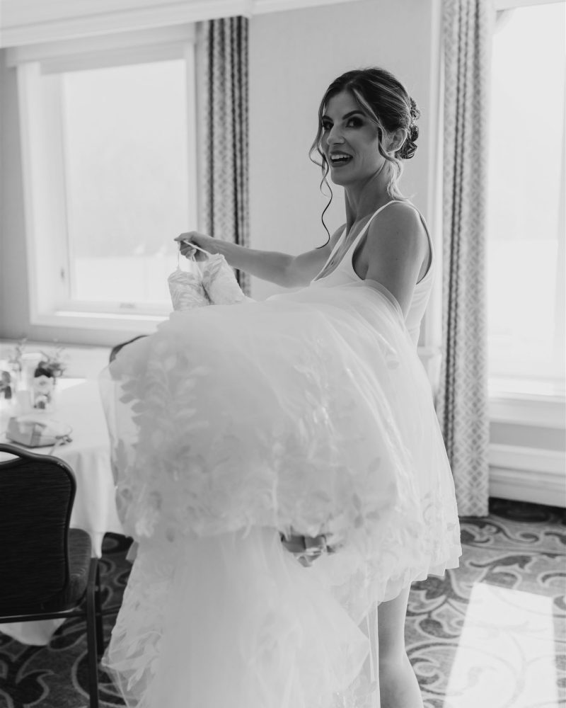 A smiling bride in a tank top holds up her wedding dress in a bright, elegant room with large windows and patterned carpet—capturing the excitement of a Banff wedding. The image is in black and white.
