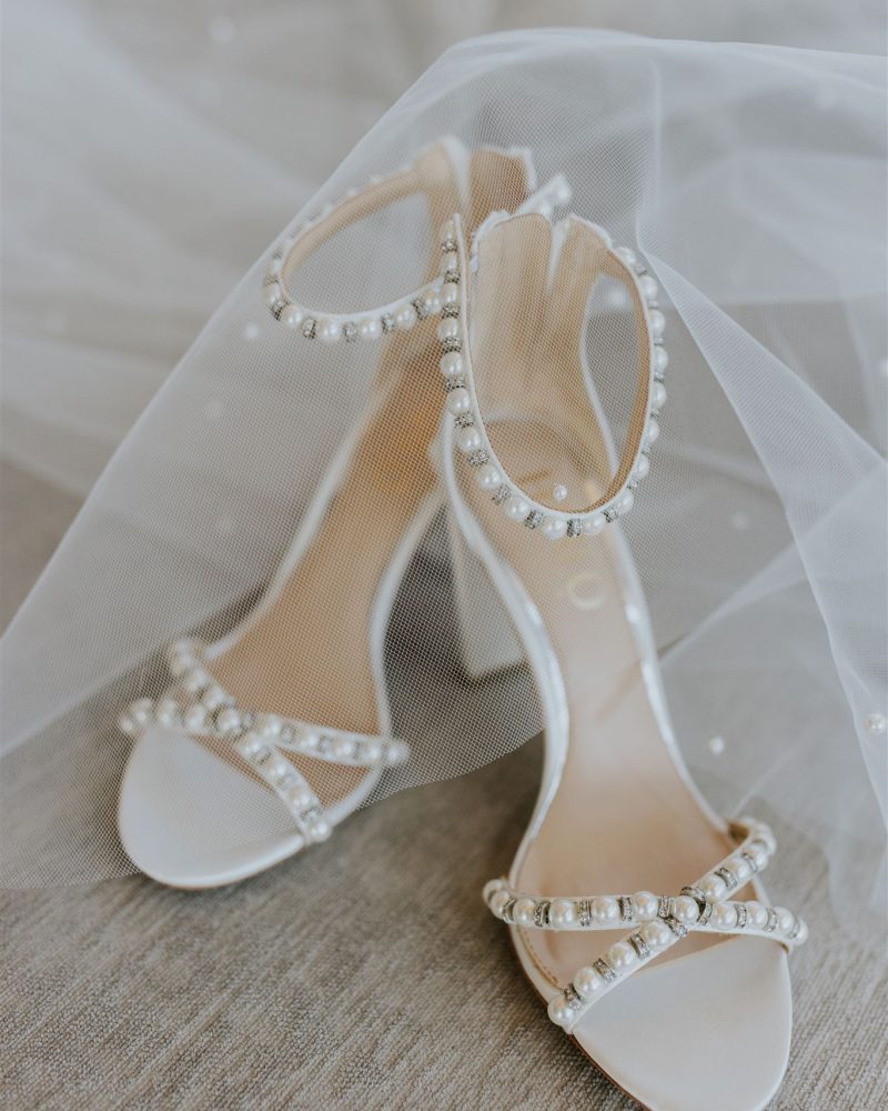 A pair of elegant high-heeled sandals adorned with pearls, perfect for a Calgary elopement, displayed on light fabric and partially covered by a sheer white veil.