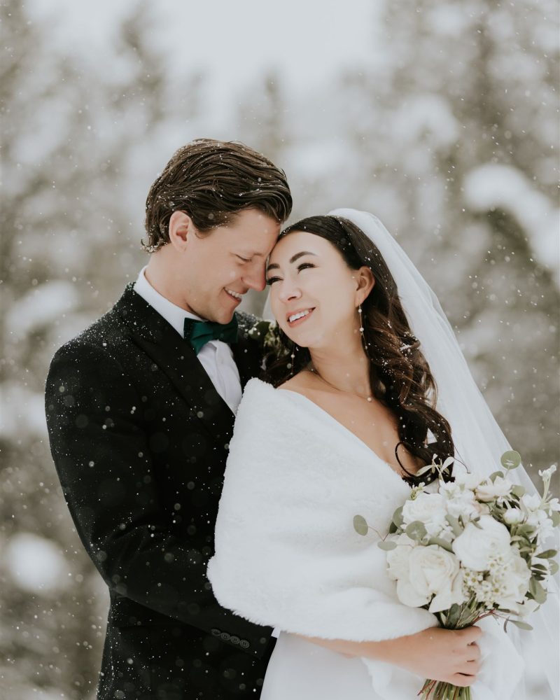 A bride and groom stand close together in the snow, smiling warmly on their Calgary elopement. The bride wears a white shawl and holds a bouquet, while the groom is in a black suit. Snow gently falls around them with trees in the background.
