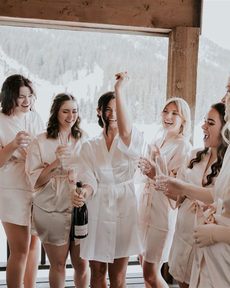 A group of seven women in matching light pink robes stand on a balcony in Banff, smiling and celebrating with champagne and glasses, surrounded by snowy mountain scenery.