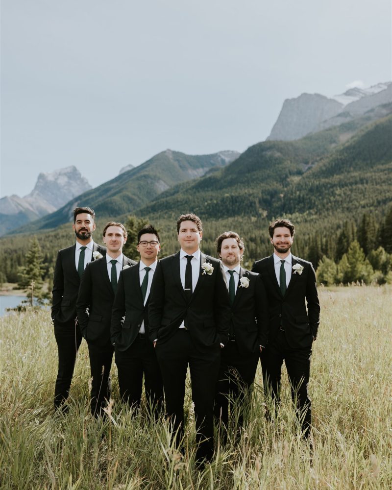 Six men in matching black suits and ties stand together in a grassy field near Canmore, with mountains and trees in the background, smiling and posing for a group photo by a photographer on a clear day.