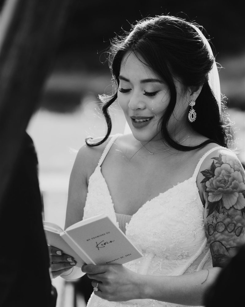 A bride in a white dress with a floral tattoo on her arm reads from a booklet during an outdoor wedding ceremony. Captured by a Calgary photographer, she smiles softly in this black and white photo.