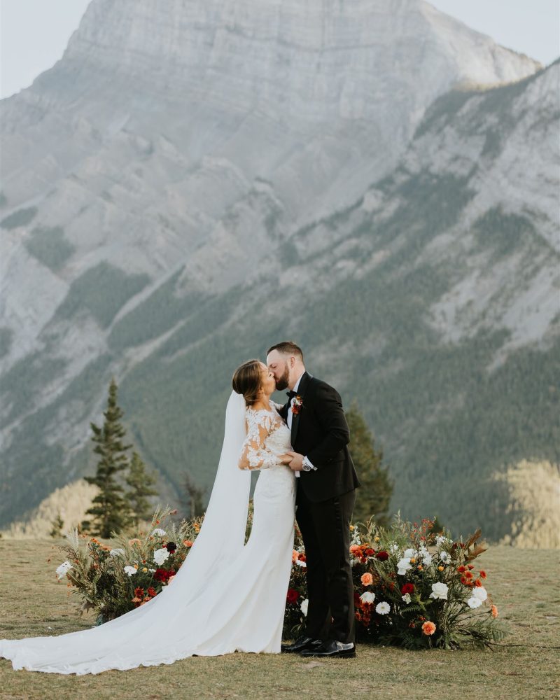 A bride and groom share a kiss outdoors in front of a mountain backdrop in Canmore, surrounded by colorful flower arrangements—a perfect setting for an intimate wedding. The bride wears a long white dress and veil; the groom is in a black suit.