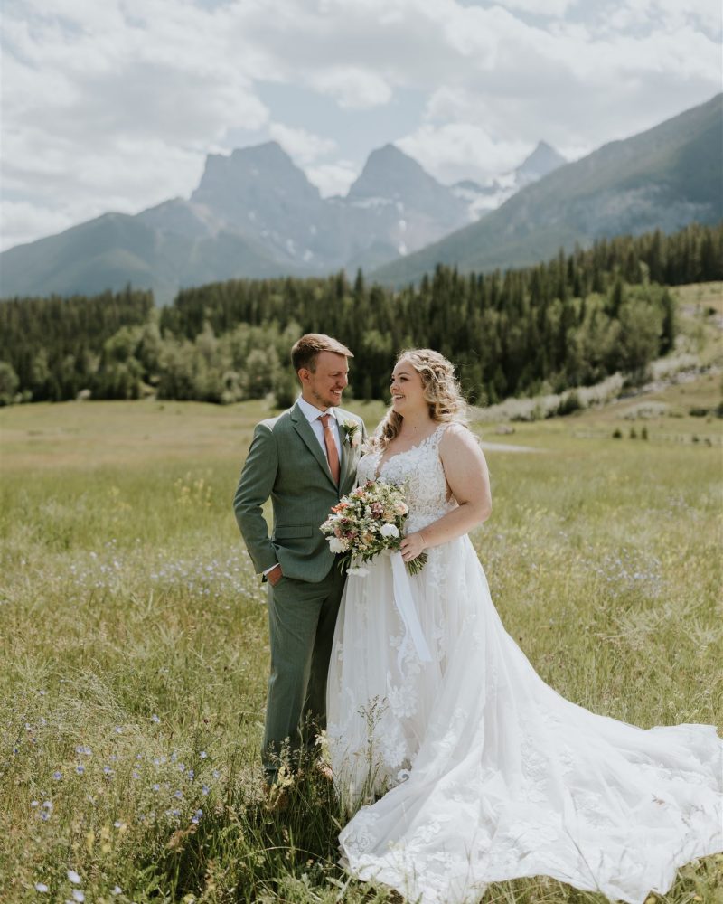 A bride in a white dress and a groom in a light green suit stand together smiling in a grassy field, celebrating their Canmore elopement with mountains and trees in the background under a partly cloudy sky.