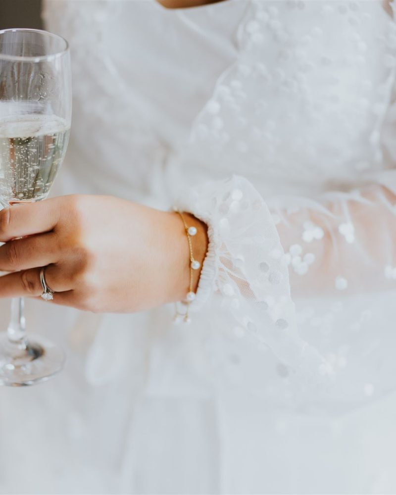 A person in a white, sheer, polka-dot dress holds a glass of sparkling wine at a Calgary wedding. A bracelet and ring are visible on their left hand. The softly blurred background adds a touch of elegance.