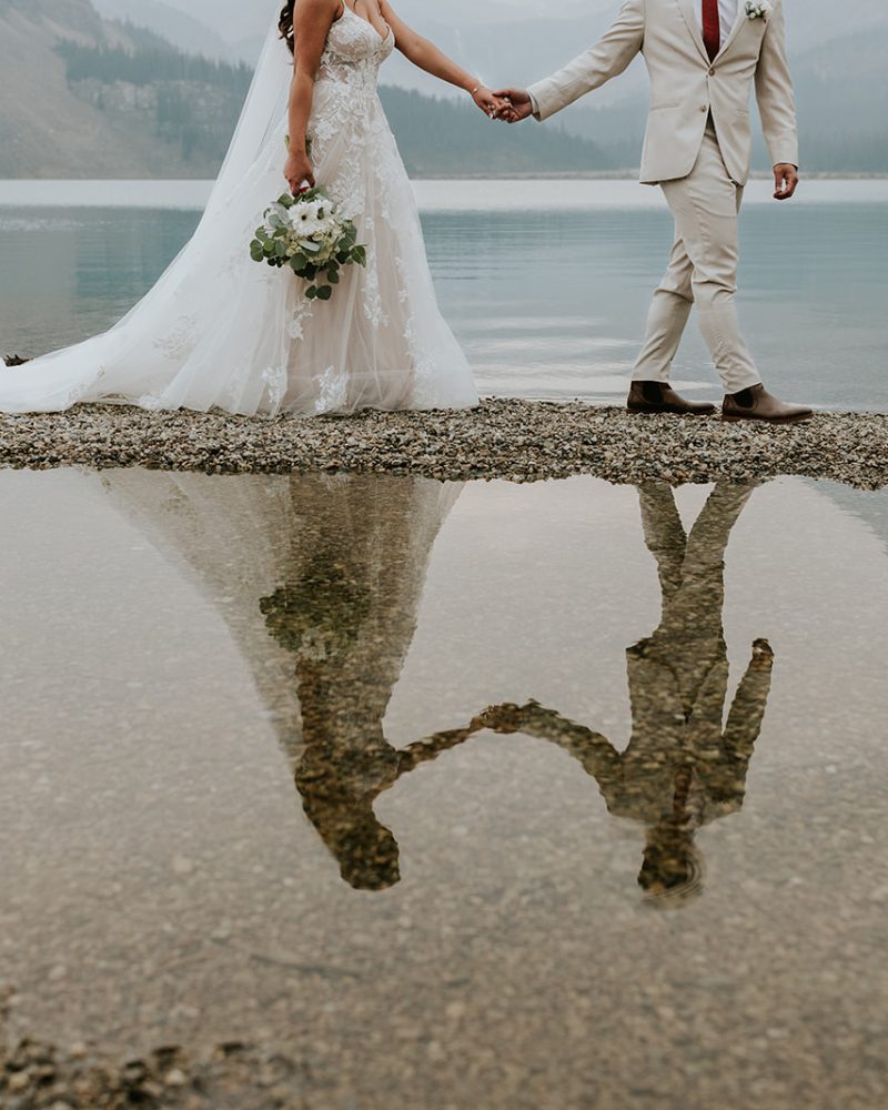 A bride and groom hold hands near a calm lake in Banff, their reflection visible in a puddle on rocky ground. The bride wears a white dress and veil; the groom is in a light suit with a red tie. Misty mountains create the perfect elopement backdrop.