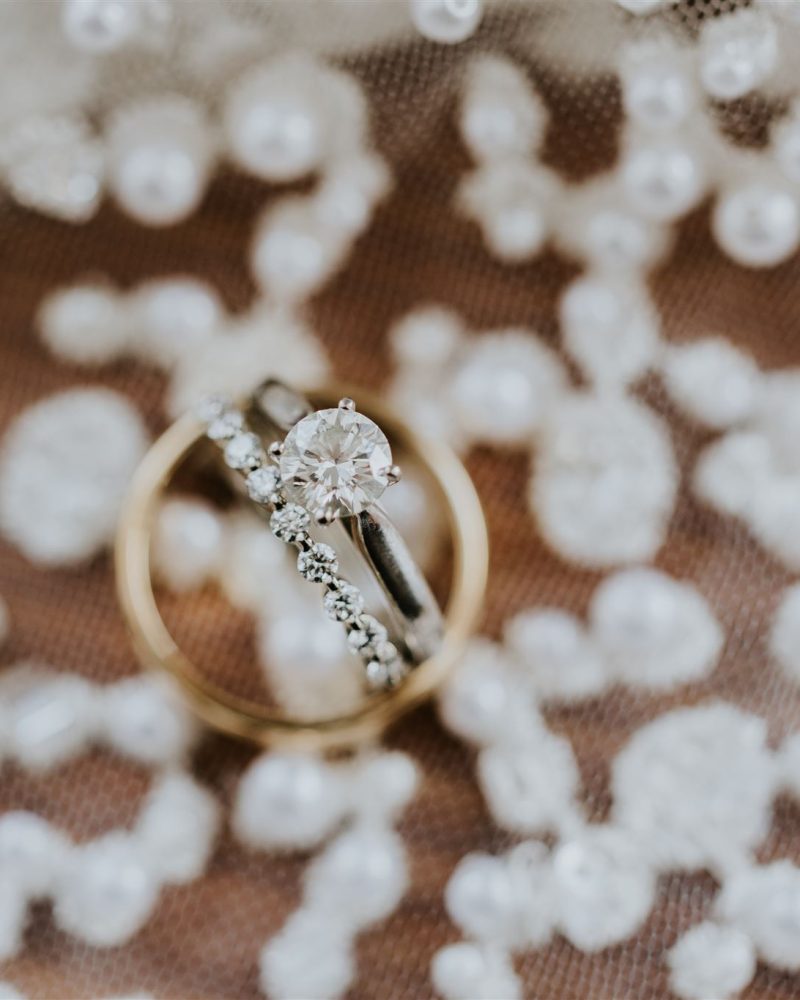Close-up of a diamond engagement ring and wedding band resting on intricate white lace with pearl details. The rings, perfect for an intimate Banff elopement, are positioned together to highlight their sparkling stones and elegant design.