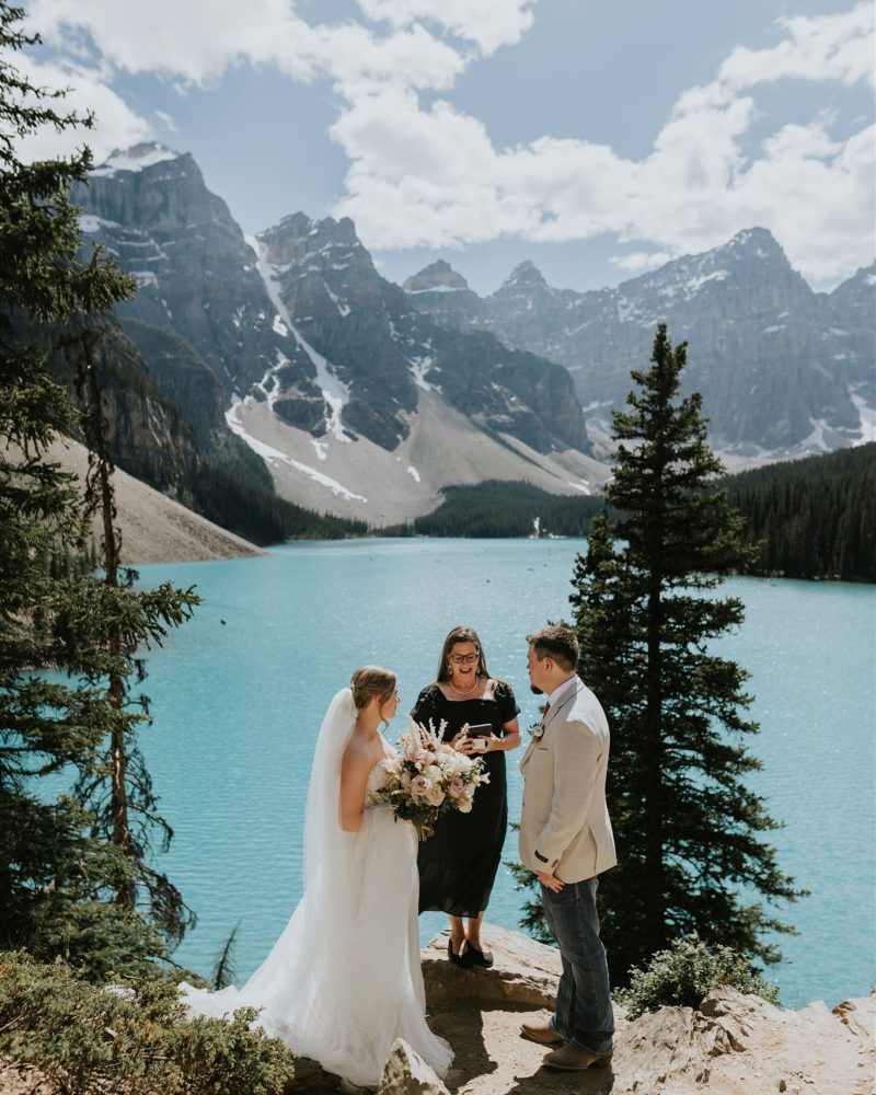 A bride and groom stand with an officiant on a rocky ledge overlooking a turquoise lake and snowy mountains near Canmore, surrounded by pine trees, as their photographer captures this stunning outdoor wedding ceremony.