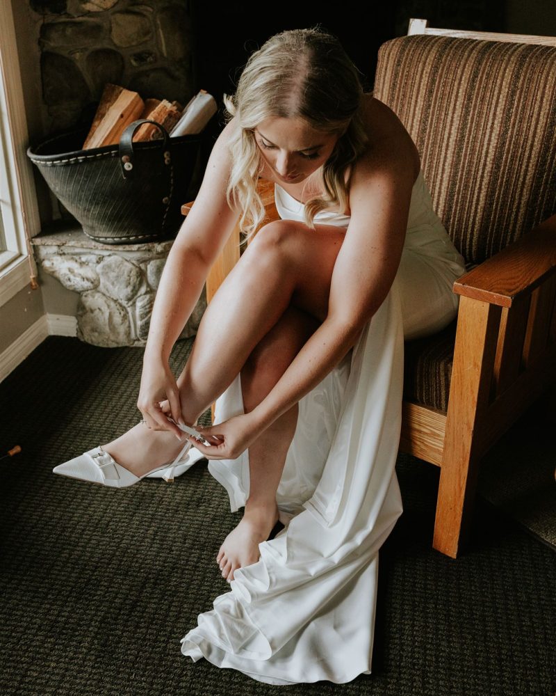 A bride with long blonde hair sits on a striped armchair in Banff, wearing a white wedding dress as she puts on a white shoe. A stone fireplace and basket of firewood are in the background.
