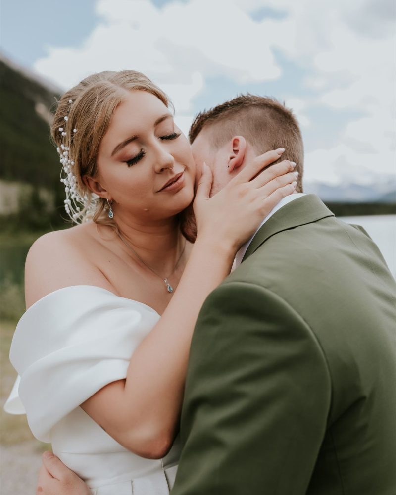 A bride in an off-shoulder white dress gently embraces her groom in a green suit. Captured by a Calgary wedding photographer, they share a tender moment by a lake with mountain views in the background.