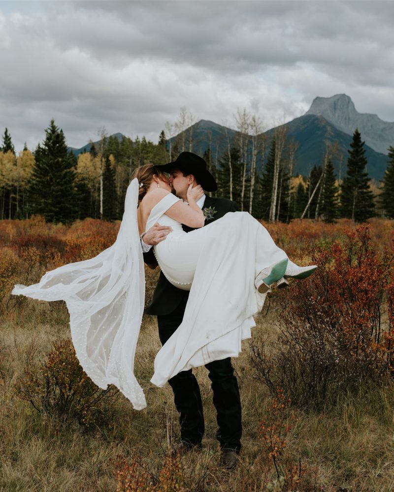 A Calgary wedding photographer captures a groom in a black hat lifting and kissing his bride in a white dress, set in a scenic autumn field with mountains, evergreen trees, and cloudy skies in the background.