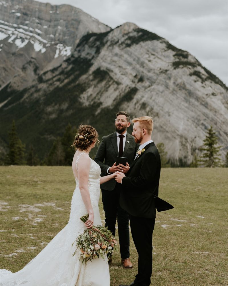 A bride and groom stand facing each other, holding hands during an outdoor Canmore wedding ceremony, with an officiant between them. A photographer captures the moment against snow-capped mountains and cloudy skies.