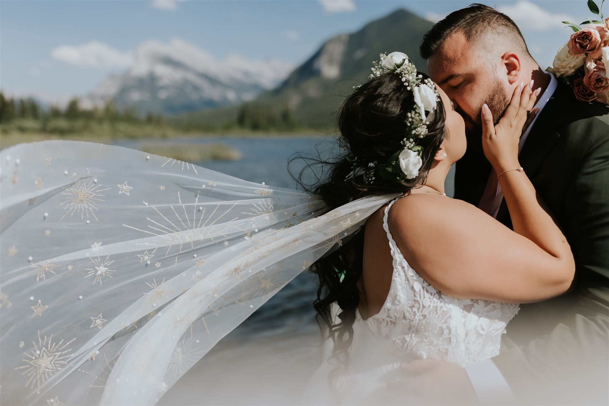 A bride and groom share a kiss outdoors by a lake with mountains in the background during their Banff elopement. The bride’s white veil, adorned with gold star patterns, flows in the wind as she wears a floral crown.