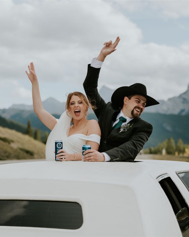 A joyful bride and groom, holding drinks, lean out of the sunroof of a white limousine. Smiling and raising their arms, they celebrate amidst the stunning mountains and trees near Canmore under a cloudy sky.