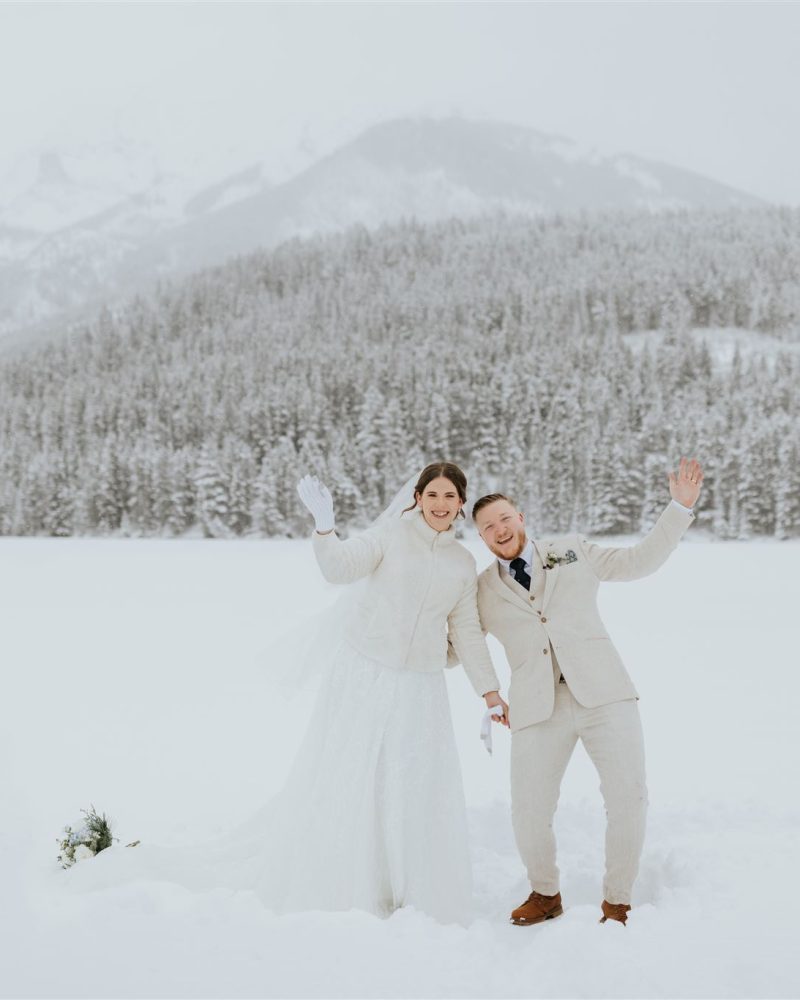 A joyful bride and groom dressed in white winter attire stand in a snowy landscape near Banff, waving and smiling in front of a snow-covered forest and mountain backdrop.