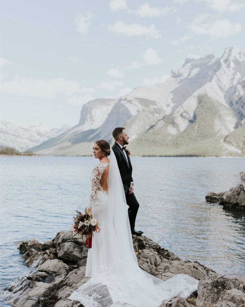 A bride in a lace dress and veil stands holding a bouquet next to a groom in a suit, both on a rocky shore by a lake at Banff, with tall mountains in the background—an intimate elopement under a partly cloudy sky.