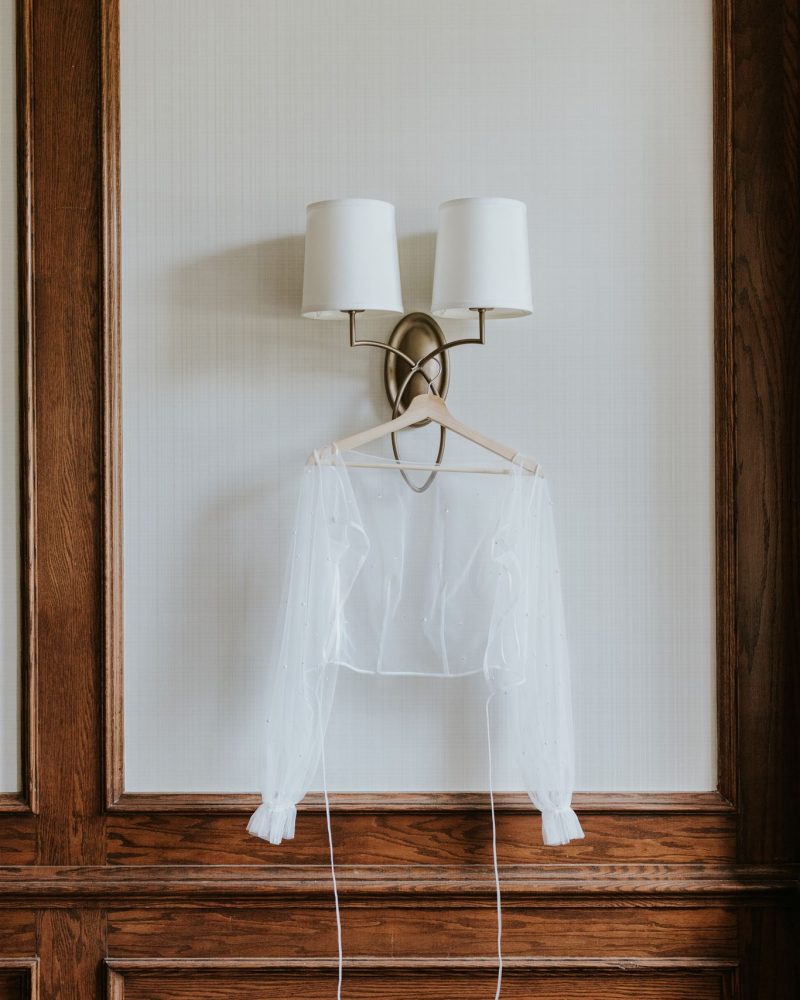 A sheer, long-sleeve white blouse hangs on a wooden hanger, perfect for a wedding elopement, hooked to a wall sconce with two white lampshades, set against wood-paneled walls and a white textured backdrop.