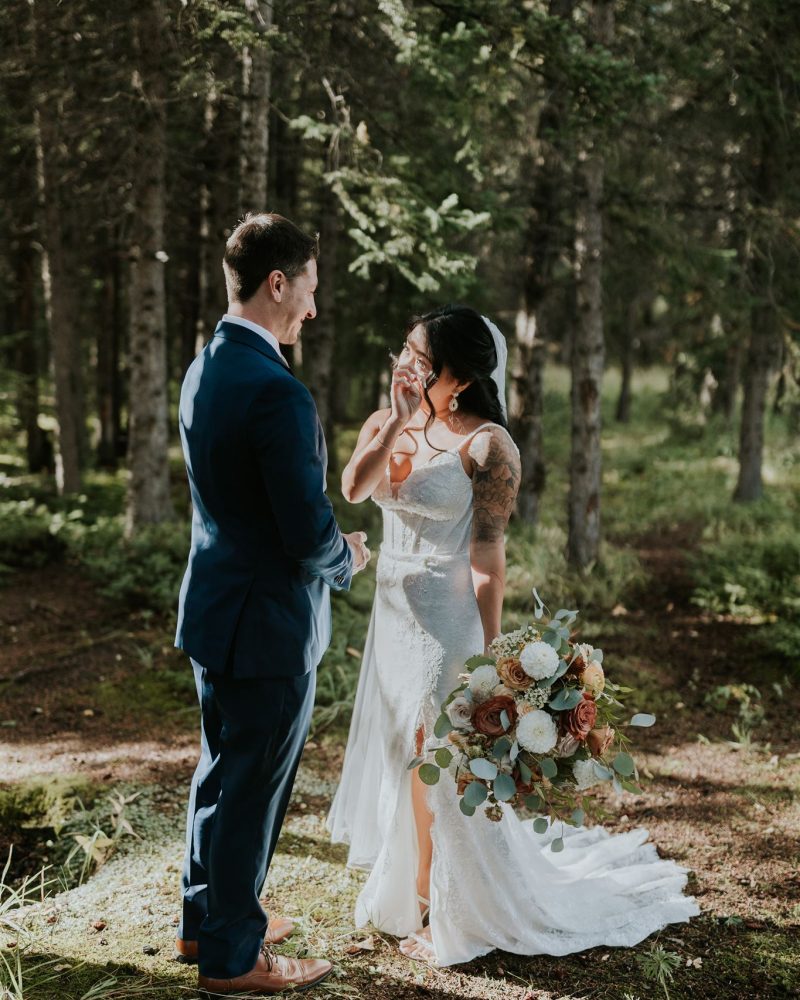 A bride in a white dress holding a bouquet wipes tears from her face, smiling at a groom in a blue suit as they stand together in a sunlit Canmore forest clearing, perfectly captured by their wedding photographer.