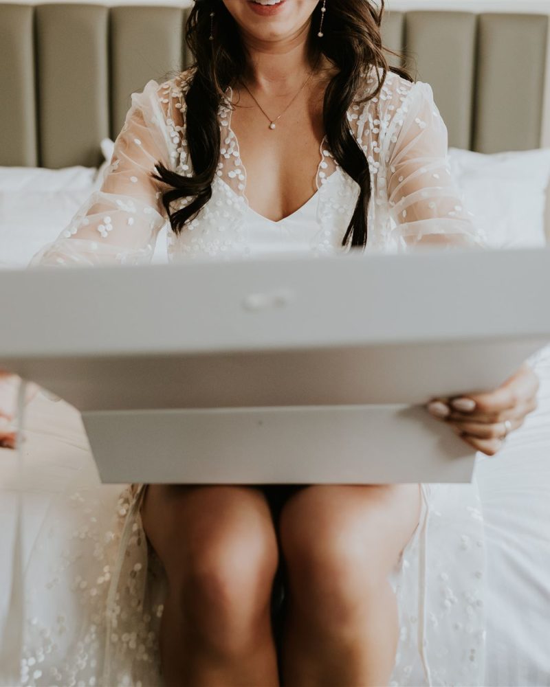 A woman in a white, sheer robe sits on a bed in a cozy Calgary bedroom, smiling as she opens a large white box in her lap. The room features neutral decor with lamps on either side of the bed.