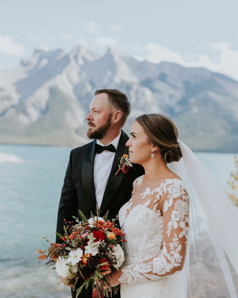 A bride in a long-sleeve lace wedding dress and veil stands beside a groom in a black tuxedo by a lake, both gazing thoughtfully ahead, with mountains and blue sky in the background—a beautiful Canmore elopement. The bride holds a floral bouquet.