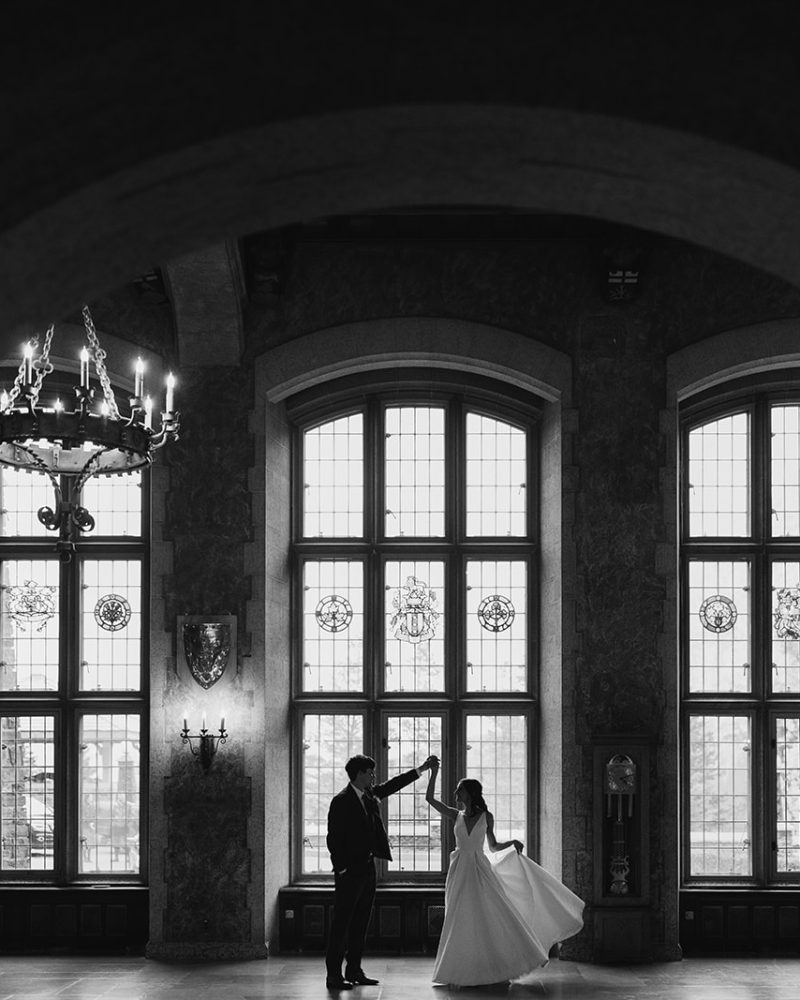 A bride and groom dance in an elegant, grand Calgary hall with tall arched windows and a large chandelier, framed by a dark archway in the foreground. This timeless wedding scene is beautifully captured in black and white.