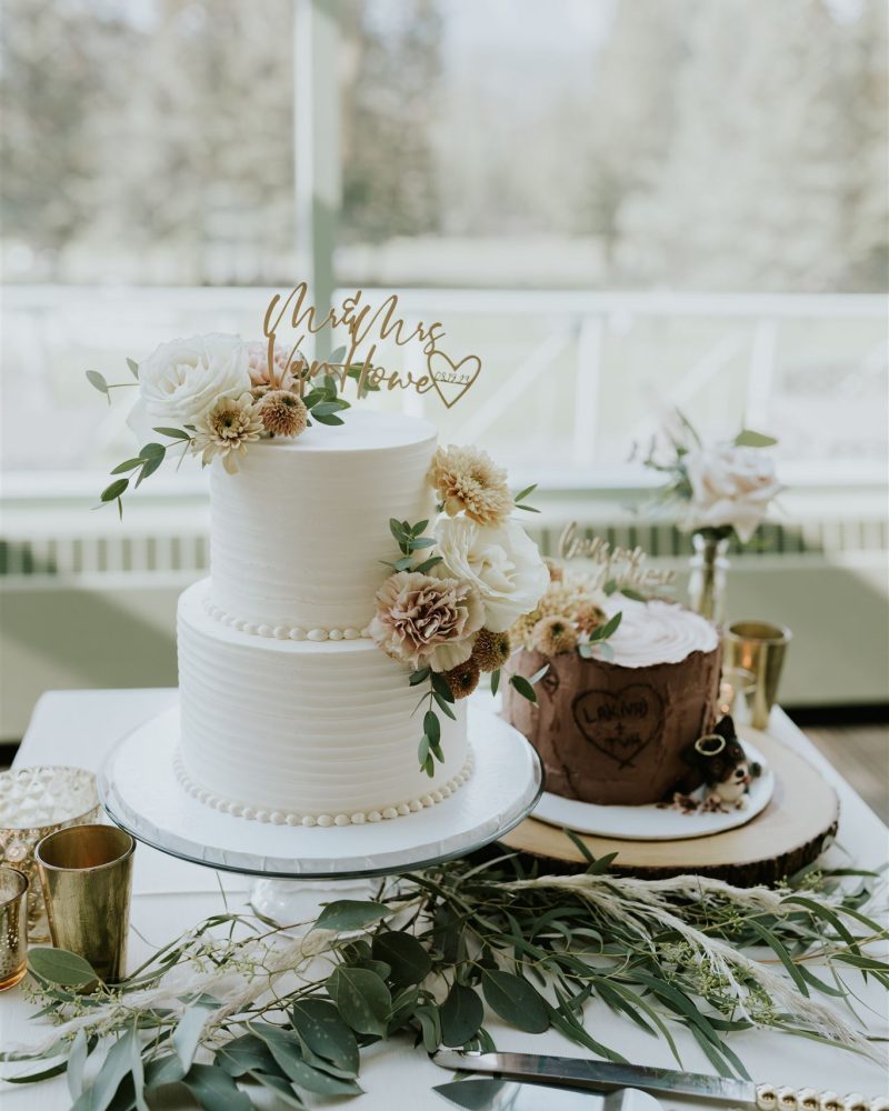 A white two-tiered wedding cake decorated with flowers and greenery sits beside a rustic tree stump cake. Both are on a table adorned with greenery, gold cups, and cutlery at a beautiful Banff or Calgary celebration.