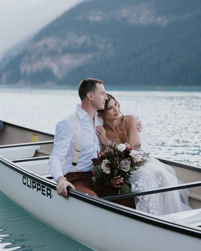 A Calgary wedding photographer captures a bride in a white dress and groom in suspenders sitting closely together in a canoe on a calm mountain lake, the bride holding a bouquet as forested mountains frame the romantic scene.