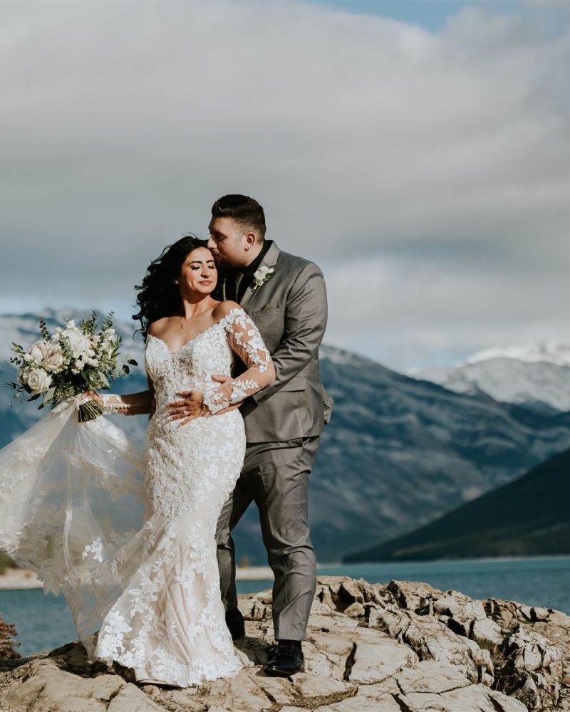 A bride in a white lace gown holds a bouquet while her groom, in a gray suit, embraces and kisses her forehead. They celebrate their elopement on rocky terrain beside a lake with Banff mountains and a cloudy sky in the background.