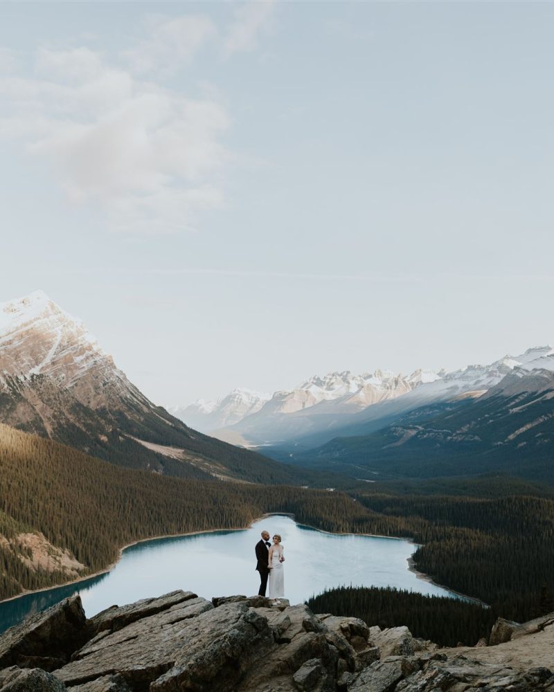 A couple stands on rocky terrain overlooking a vast mountain landscape near Calgary, with a winding blue lake below—an idyllic scene for a wedding photographer, surrounded by snow-capped peaks and dense evergreen forests under a clear sky.