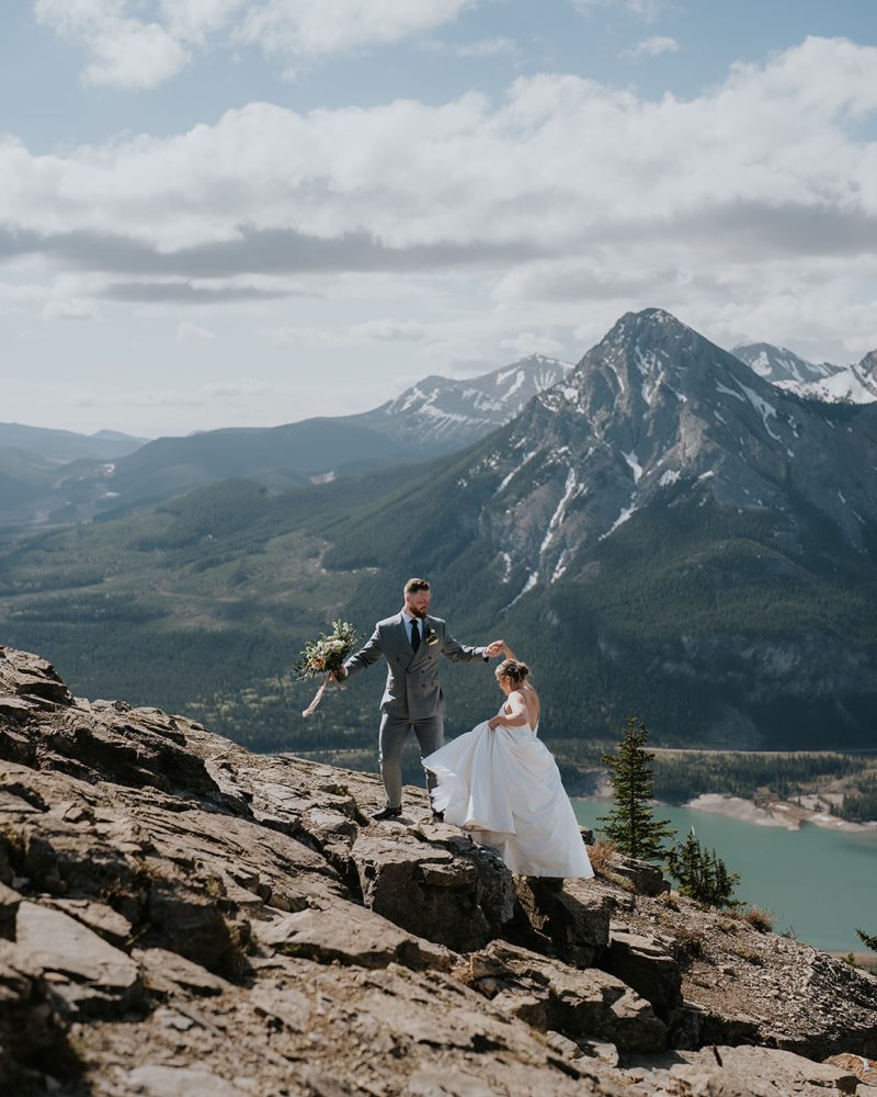 A couple in wedding attire stands on rocky mountain terrain, with the groom helping the bride up—an ideal elopement moment captured by a Calgary photographer, set against forested mountains, a lake, and a partly cloudy sky.