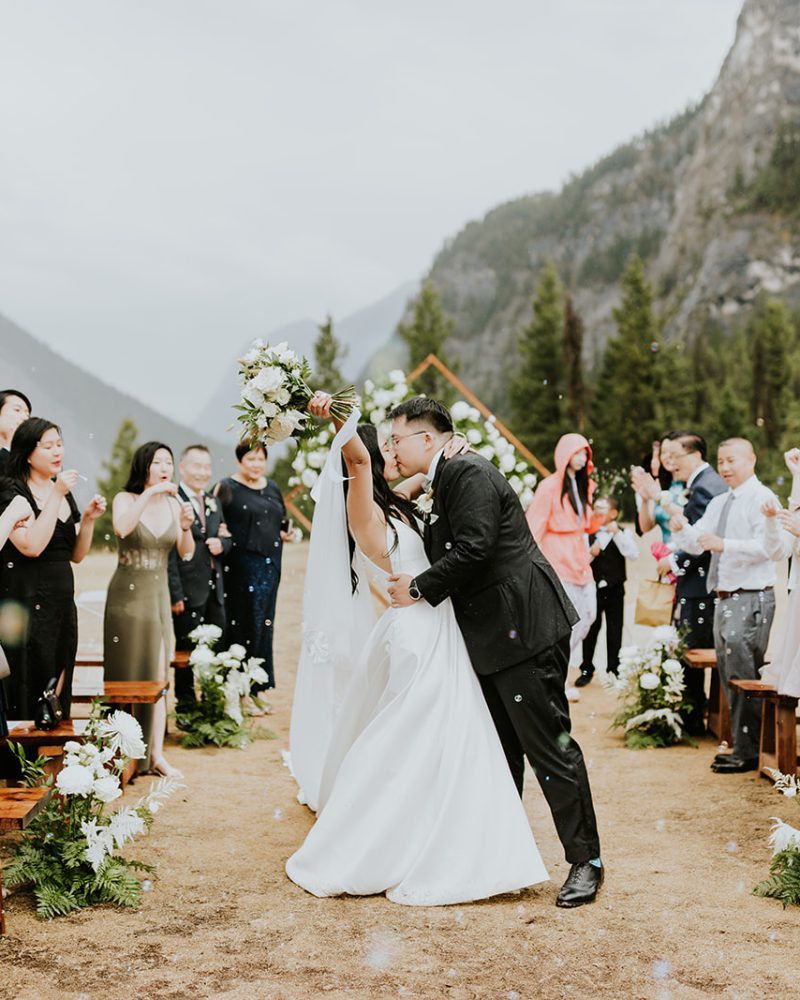 A bride and groom kiss at an outdoor Canmore wedding ceremony, surrounded by cheering guests. Mountains and pine trees form a scenic background, with floral arrangements lining the aisle.