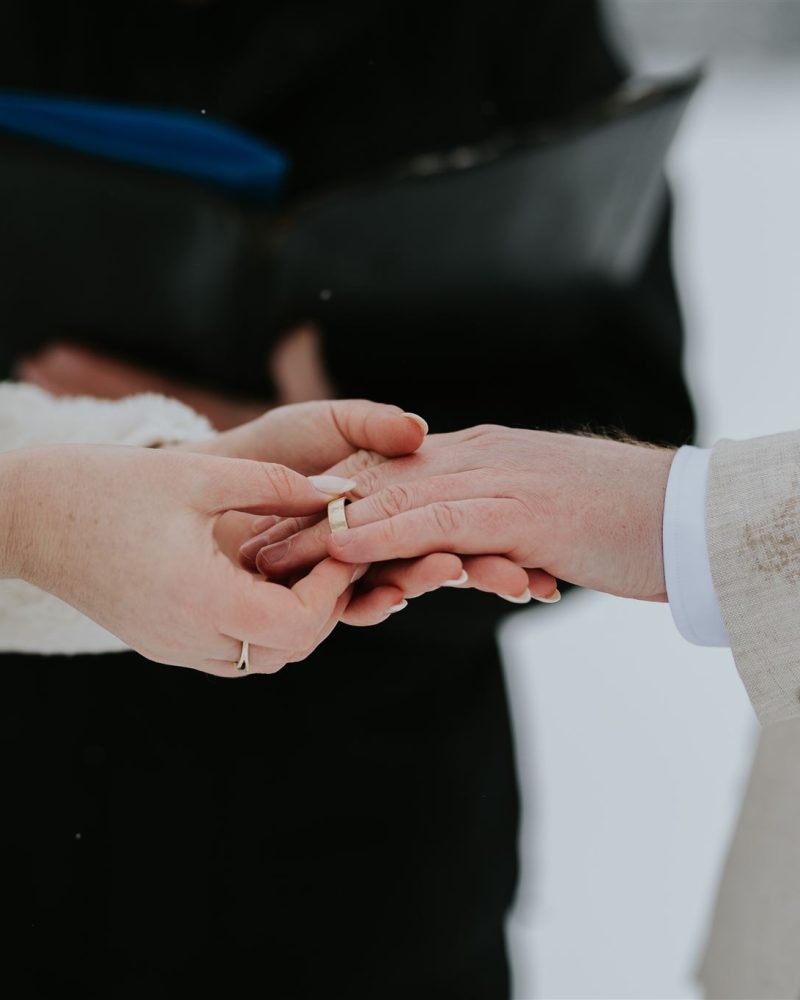 Close-up of a person in a white outfit placing a ring on another's finger, who is wearing a beige suit, during a snowy Banff wedding. The background is blurred with a third person partially visible holding a folder.