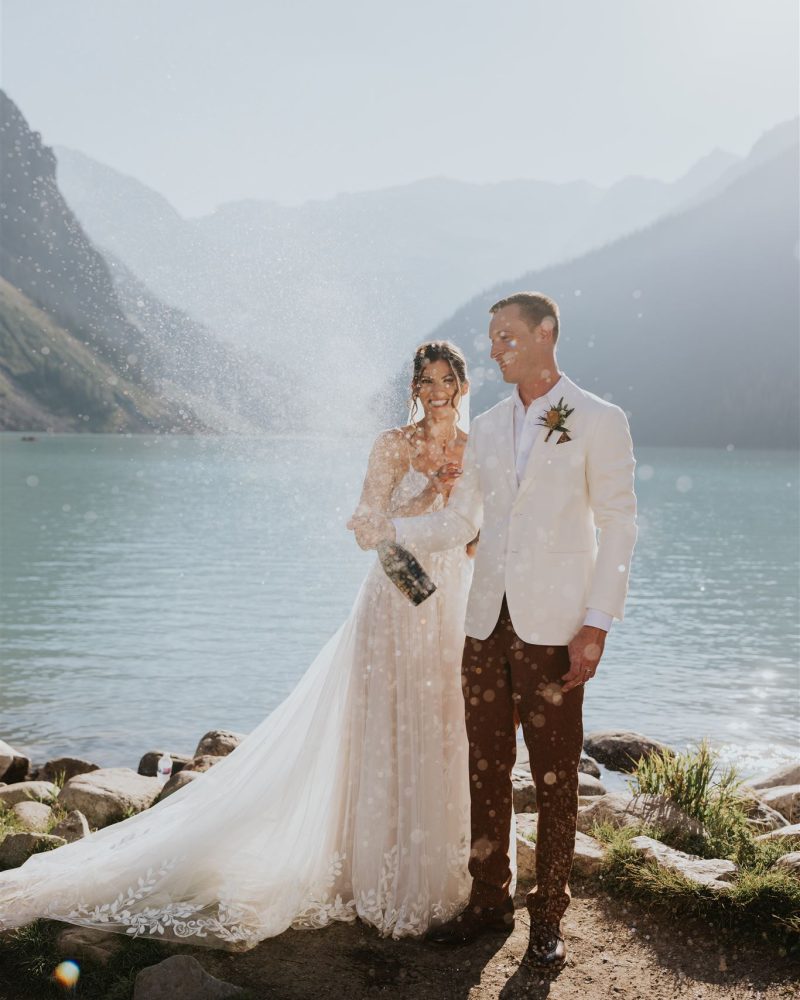 A bride and groom stand by a Banff mountain lake, the bride in a white dress and the groom in a white jacket. The bride pops a champagne bottle, spraying bubbles into the air as they smile, celebrating their elopement.
