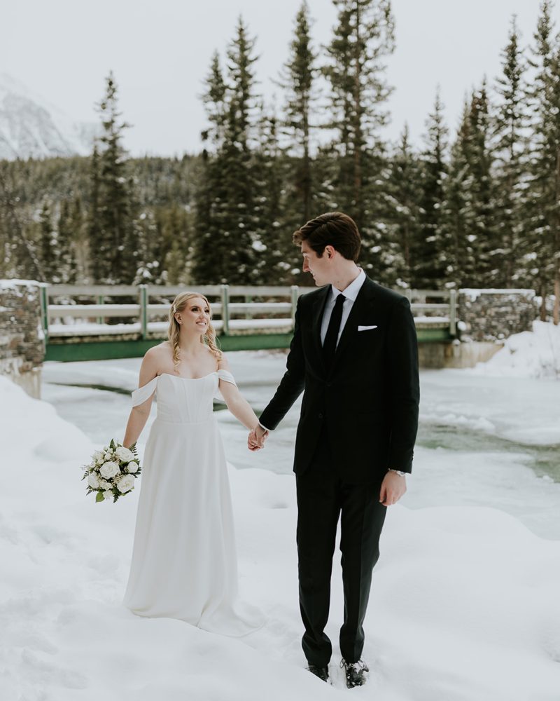 A bride in a white gown and a groom in a black suit hold hands while standing in the snow near a river, as their Calgary wedding photographer captures them smiling at each other with pine trees and a wooden bridge in the background.