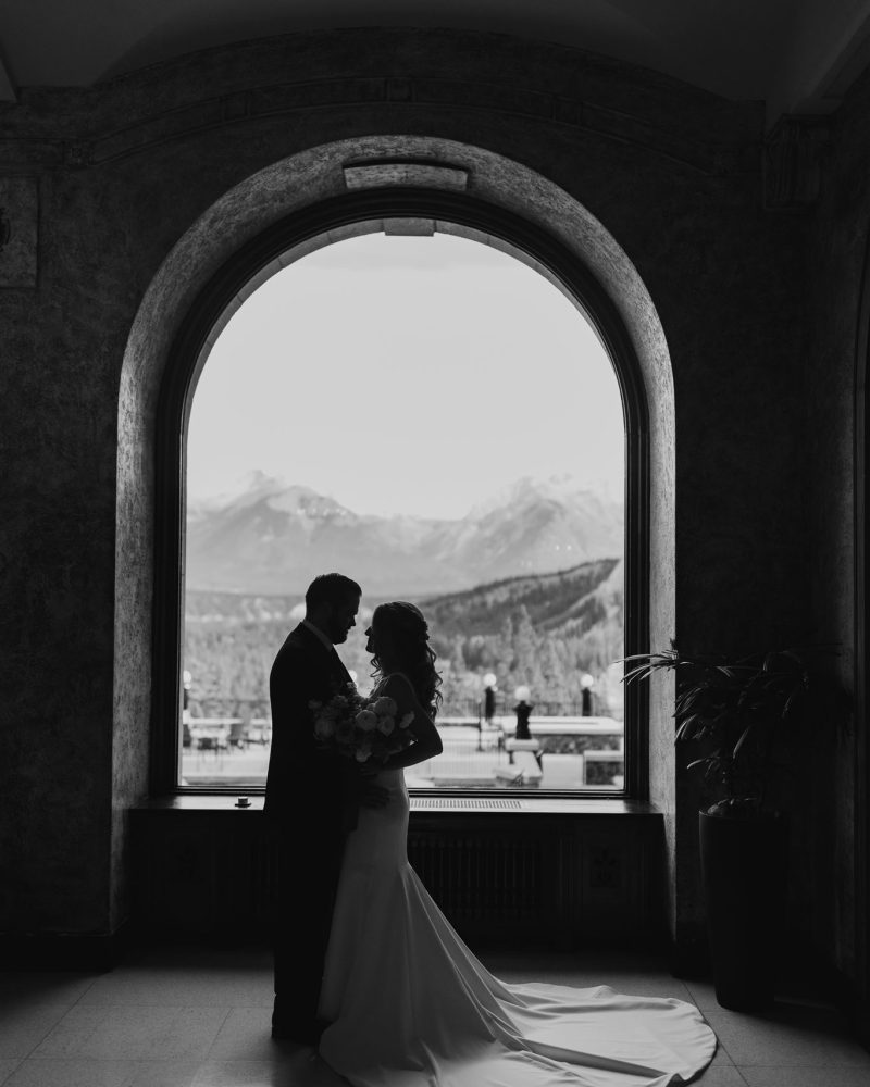 A bride and groom stand facing each other in front of a large arched window with mountains in the background, their silhouettes captured in a black-and-white photo during an intimate Calgary wedding.