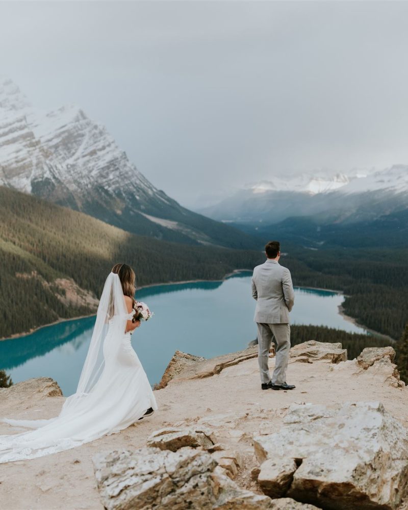 A bride in a white gown and veil walks toward her groom in a gray suit, both standing on rocky terrain overlooking a turquoise lake and snow-capped mountains—an unforgettable Canmore wedding captured by a skilled photographer.