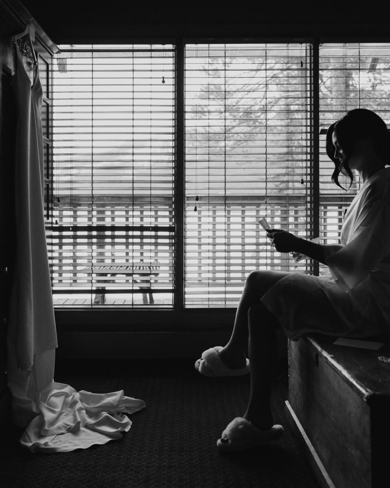 A woman in a robe sits on the edge of a bed, reading a card in a dimly lit room with large windows. A wedding dress hangs by the window as light streams through the blinds—captured perfectly by the photographer in Banff.