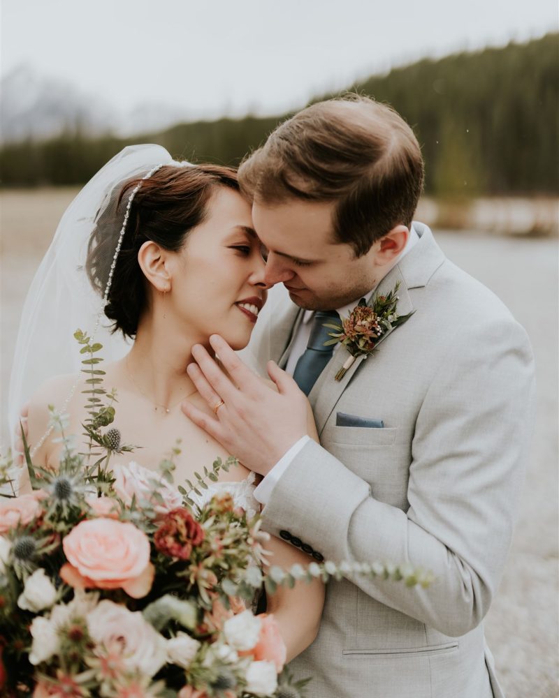 A bride and groom embrace outdoors in Canmore, smiling lovingly at each other. The bride holds a large bouquet of pink and white flowers, and both wear formal wedding attire with trees and mountains in the background.