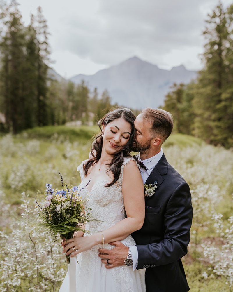 A bride and groom embrace in a lush, outdoor Canmore setting with mountains and trees in the background. The bride holds a bouquet, smiling softly with her eyes closed as the elopement photographer captures the intimate moment.